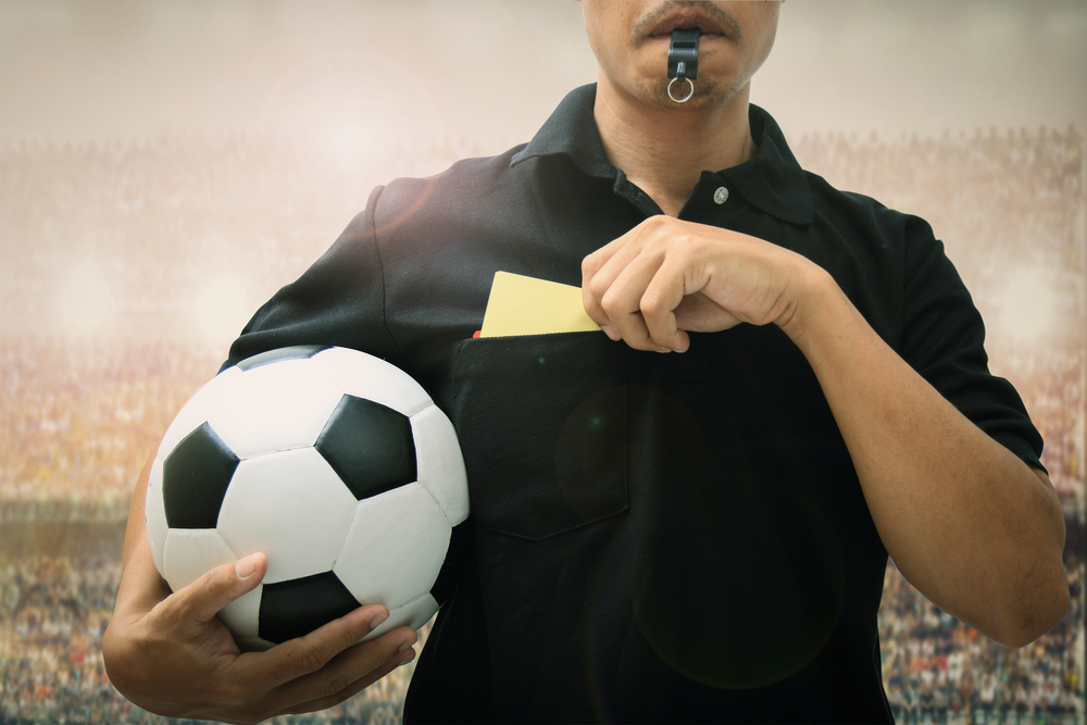 moustached soccer referee blows whistle while holding ball and yellow card